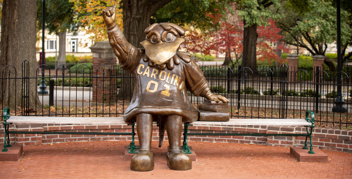 铜质公鸡雕像坐在长凳上，他的手放在一堆铜质书籍上