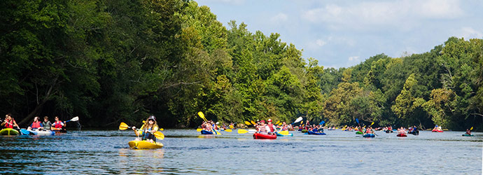 一条河上满是划着五颜六色的皮划艇的人，河岸上树木繁茂。