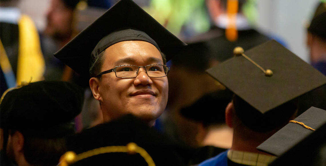 一名戴着毕业典礼帽的研究生对着镜头微笑，而其他戴着毕业典礼帽的学生则背对着他站在周围。