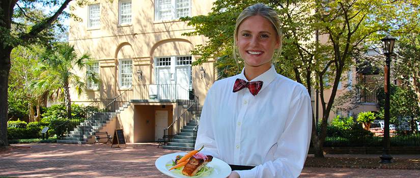 一名酒店管理专业的女学生站在历史悠久的麦卡琴大厦前的马蹄铁上。她端着一盘看起来很美味的饭菜。