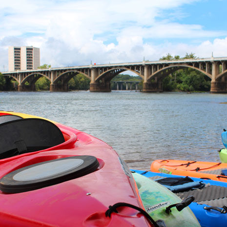 红色、橙色、蓝色和绿色的皮艇漂浮在河边，背景是一座桥梁和摩天大楼。