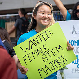 在格林街的学生组织博览会上，一名女学生举着一个牌子，上面写着“招聘STEM专业的女性”。