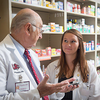 一个学生和一个教授站在一个药品货架前谈话。