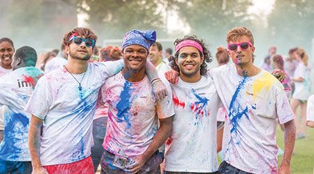 四名学生穿着溅有彩色粉笔的白衬衫，双臂环抱着对方，在一场彩色跑步中摆出姿势。