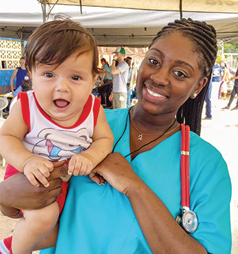 希尔梅尔·麦卡洛穿着蓝色的手术服和红色的听诊器，抱着一个婴儿，对着镜头微笑。