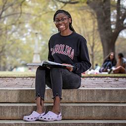Student with dark hair and a black "Carolina" sweatshirt on sitting on the horseshoe smiling.