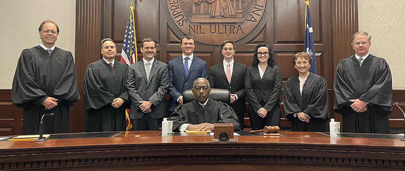 模拟法庭团队的成员被南卡罗来纳州最高法院的成员包围，站在最高法院的长凳后面，背景是最高法院的印章。