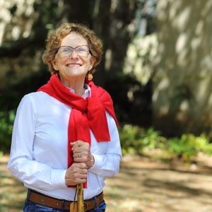 一名女子戴着眼镜，身穿白色衬衫，肩上系着红色毛衣，手里拿着一根以树叶为背景的手杖