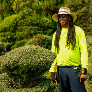 一名男子身穿绿色衬衫，戴着太阳镜，站在珍珠弗赖亚园艺花园的灌木丛旁