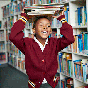 一个穿着红色毛衣的学生微笑着穿过图书馆，她的头上顶着一堆书。